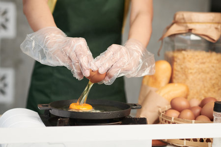 Close-up image of female cook wearing protective gloves when cracking eggs over frying panの写真素材