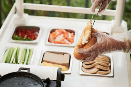 Hands of vendor wearing protective gloves when making sandwich with fried eggs, vegetables and meat for customerの写真素材
