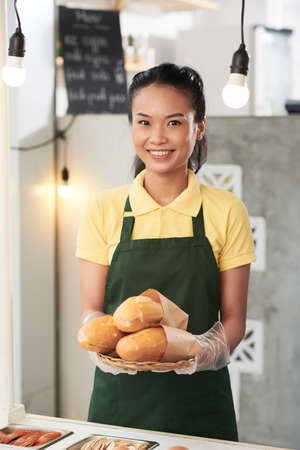 Portrait of happy smiling young woman in protective gloves working as vendor and offering plate of sandwichesの写真素材