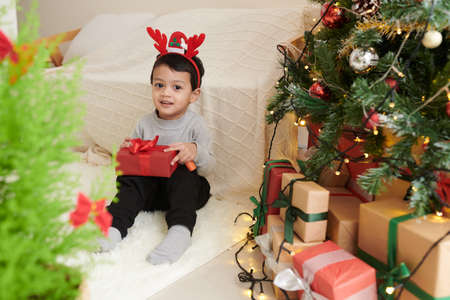 Cute little boy wearing reindeer antlers headband when sitting next to decorated Christmas tree with present in handsの写真素材