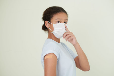 Portrait of teenage girl in medical mask having adhesive plaster over her covid-19 vaccine injection...