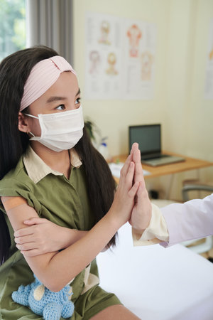 Little girl in medical mask giving high five to general practitioner after getting vaccine in...