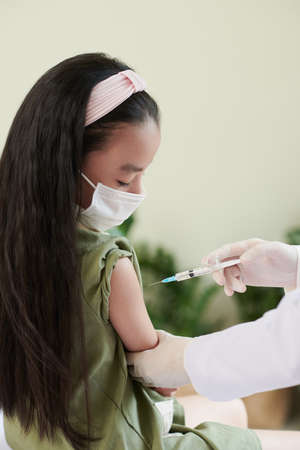 Serious little girl in medical mask looking at doctor injecting vaccine in her armの写真素材