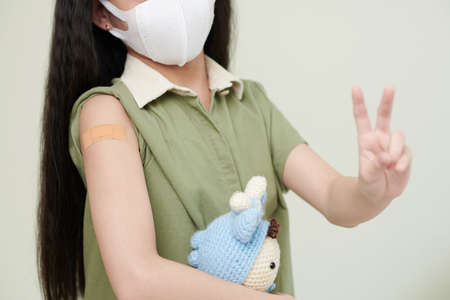 Little girl in protective mask having adhesive plaster on her shoulder and showing peace gestureの写真素材