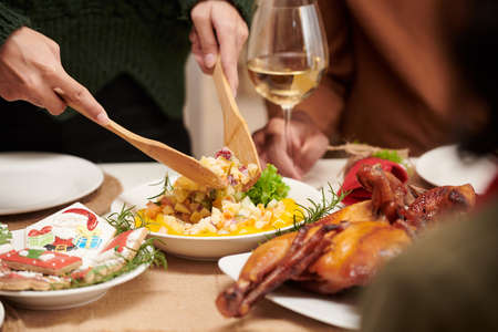 Hands of woman using wooden spoon when putting salad from main dish in her plate at family dinnerの写真素材