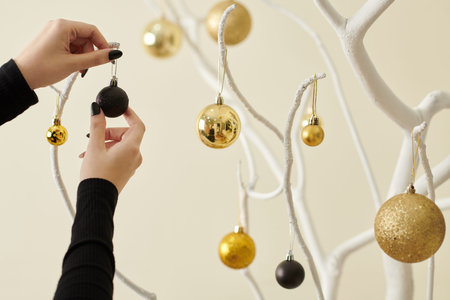 Cropped image of woman decorating bare tree branches painted white with black and golden baubles