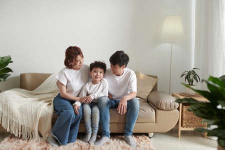 Happy parents and their smiling son in jeans sitting on sofa at homeの写真素材