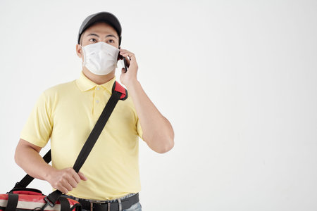 Portrait of serious courier in protective mask talking on phone with customer, isolated on white