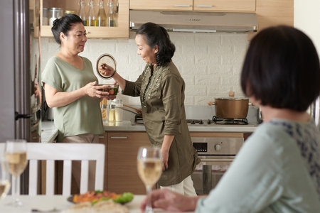Senior woman helping friend with cooking and serving dinnerの写真素材
