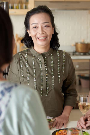 Positive senior woman standing at kitchen table and talking to friend at partyの写真素材
