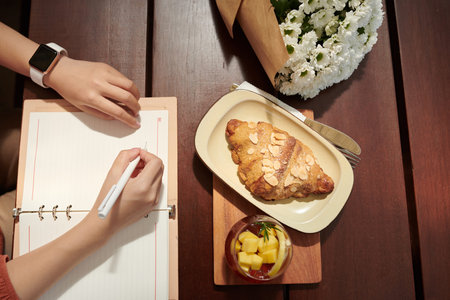 Hands of woman writing in gratitude journal when eating croissant for breakfast in cafe