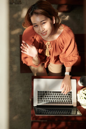 Smiling young woman coding on laptop, looking up at camera and waving with hand