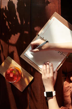 Hands of creative young woman drinking fruit cocktail and writing in planner, view from the topの写真素材