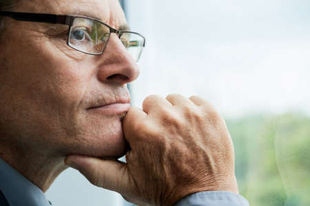 Headshot of thoughtful mature Caucasian man in glasses touching chin and contemplating cityscape through windowの写真素材