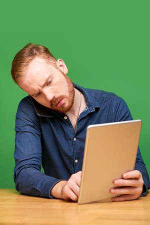 Concentrated young Caucasian man with beard sitting at wooden table and working with several devices, green backgroundの写真素材