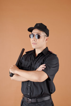 Police worker in sunglasses holding baton and crossing arms