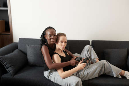 Hugging young diverse lesbian couple sitting on sofa at home and playing videogameの写真素材