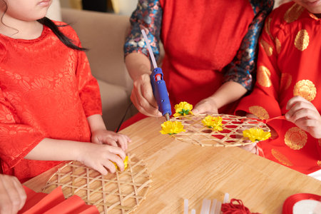 Family members using glue gun to stick yellow artificial flowers to wall decoration for tet...