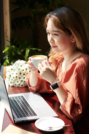 Portrait of young woman enjoying cup of morning coffee and reading news in laptop screen when...