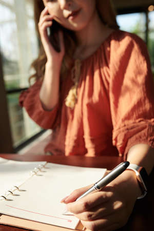 Young businesswoman talking on phone with customer or coworker and writing down important informationの写真素材