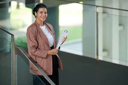 Portrait of smiling young female entrepreneur standing with clipboard on stairs in officeの写真素材