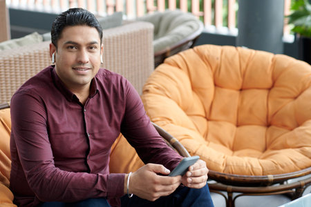 Portrait of smiling businessman in maroon shirt sitting in armchair in lounge area and wearing earbuds when calling coworkerの写真素材