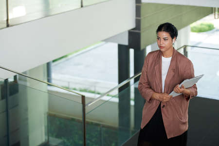 Serious young businesswoman with earbuds in ears making phone call when carrying document to upper floorの写真素材