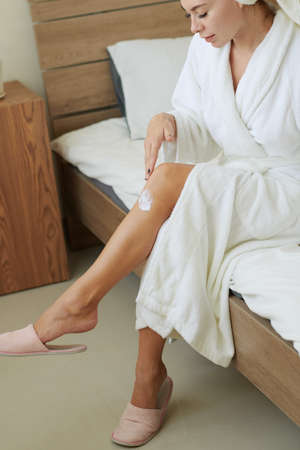 Young woman in bathrobe sitting on bed and applying rich bod creamの写真素材