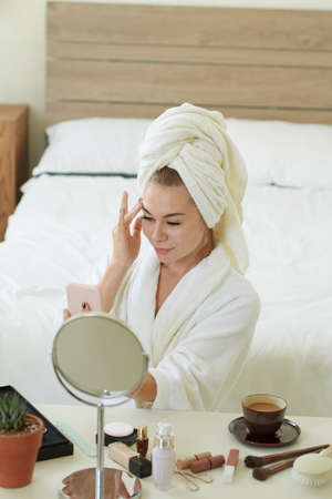 Young woman in bathrobe sitting at vanity with cup of morning coffee and applying makeupの写真素材