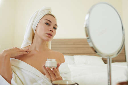 Young woman in bathrobe applying body lotion on chest in front of mirror in bedroomの写真素材