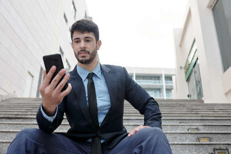 Young businessman sitting on steps outdoors and reading text messages from colleagueの写真素材