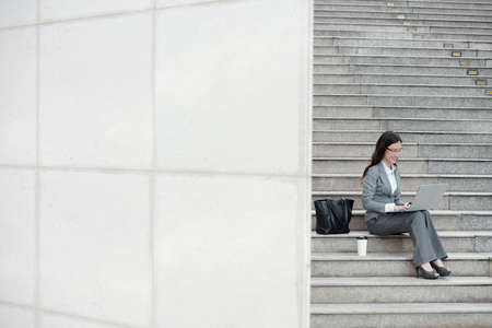 Positive young female entrepreneur sitting on steps next to her bag and cup of coffee and working on laptopの写真素材