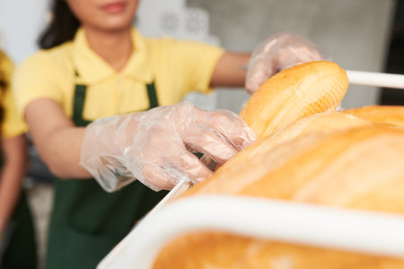Woman in disposable gloves taking fresh bun from shelf to make sandwich forの写真素材
