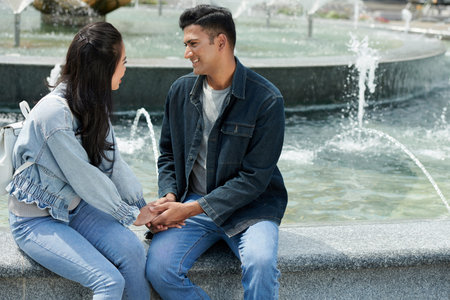 Cheerful young man and woman sitting at fountain, holding hands, talking and looking at each otherの写真素材
