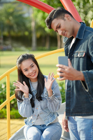 Pretty happy young woman smiling when boyfriend photographing her on smartphone