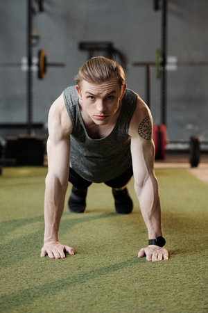 Determined fit young sportsman standing in plank position on gym floor and looking at camera