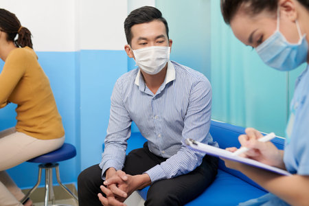 Patient in protective mask talking to medical nurse and explaining his complains and disease symptomsの写真素材