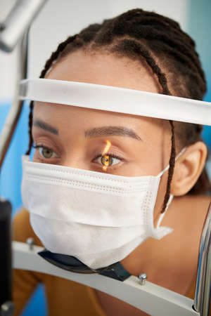 Female patient in medical mask having her eyes examined with slit lamp, microscope that allows doctor to closely examine the eyeの写真素材