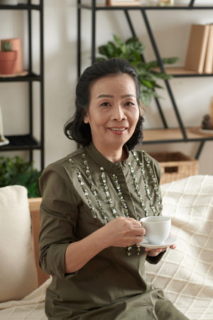 Portrait of smiling elegant senior woman sitting on sofa with up of tea in hands and looking at...