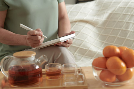 Cropped image of woman drinking tea and writing down shopping list when sitting of sofa in room