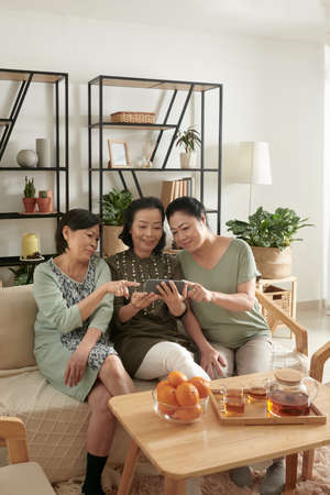 Senior woman showing photos of grandchildren to her friends when they are gathering at home for small partyの写真素材