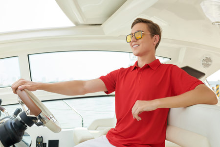 Successful confident smiling young man in red t-shirt navigating sail boat or yacht floating in sea