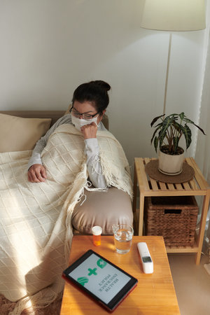 Sick woman in protective mask sitting on couch under blanket after taking medicine and filling health insurance claim formの写真素材
