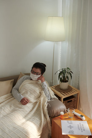 Sick senior woman in medical mask sitting on sofa under warm blanket, health insurance policy...