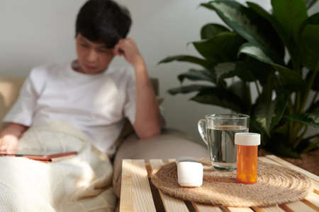 Cup of water, pills and contactless electronic thermometer on table beside sofa, sick man reading e-book or article in backgroundの写真素材