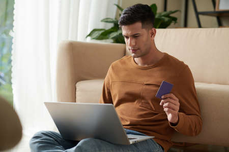 Young man sitting on the floor in the room with laptop on his knees paying purchases online using his credit cardの写真素材