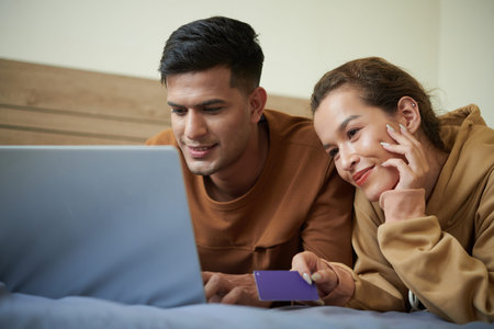 Young couple lying together on the bed in bedroom and using laptop for online delivery, they buying...