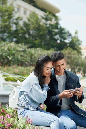 Smiling man showing funny video on smartphone to his girlfriend when they are sitting in parkの写真素材