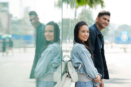 Smiling young couple waiting for bus outdoors, leaning on building and looking at roadの写真素材