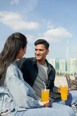 Happy young couple spending time on river embarkment, sitting on bench, talking and having soft frinksの写真素材